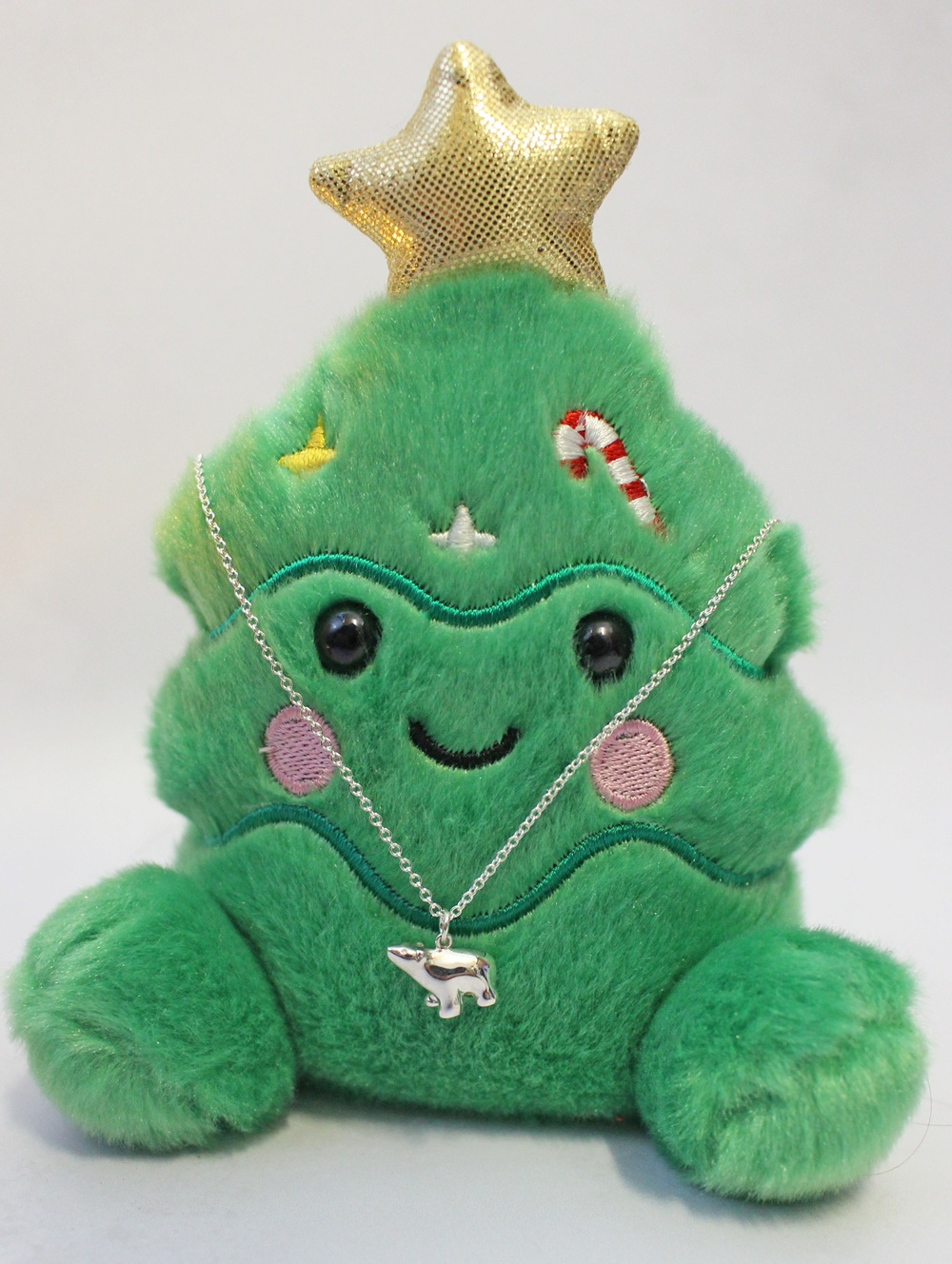 A green plush Christmas tree toy with a gold star topper, wearing a silver chain necklace with a small polar bear pendant.