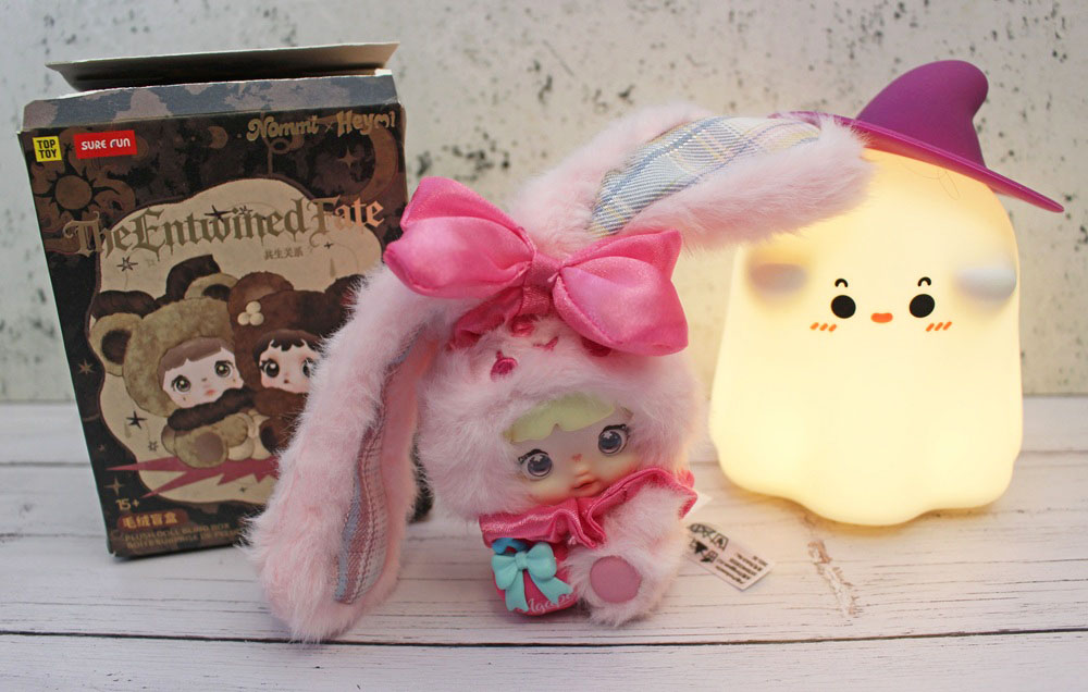 A plush toy with long pink bunny ears and a bow sits in front of its The Entwined Fate blind box and a glowing, ghost-shaped lamp wearing a witch’s hat.