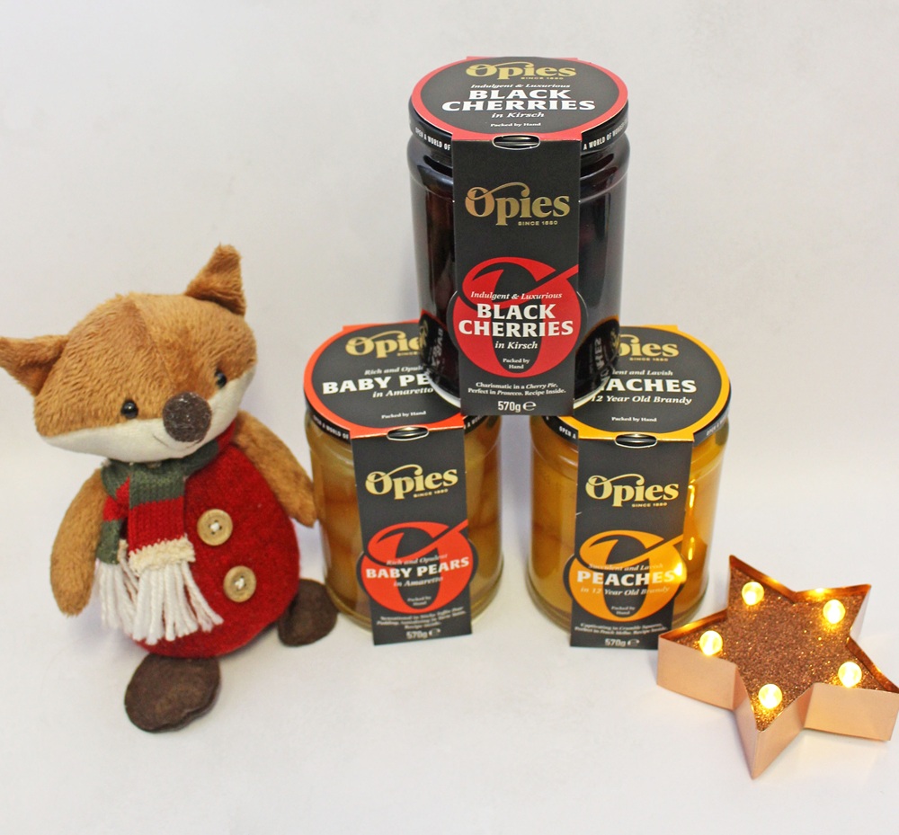Three jars of Opies fruit in brandy or liqueur—black cherries, baby pears, and peaches—arranged with a small plush fox and a lit star-shaped ornament.