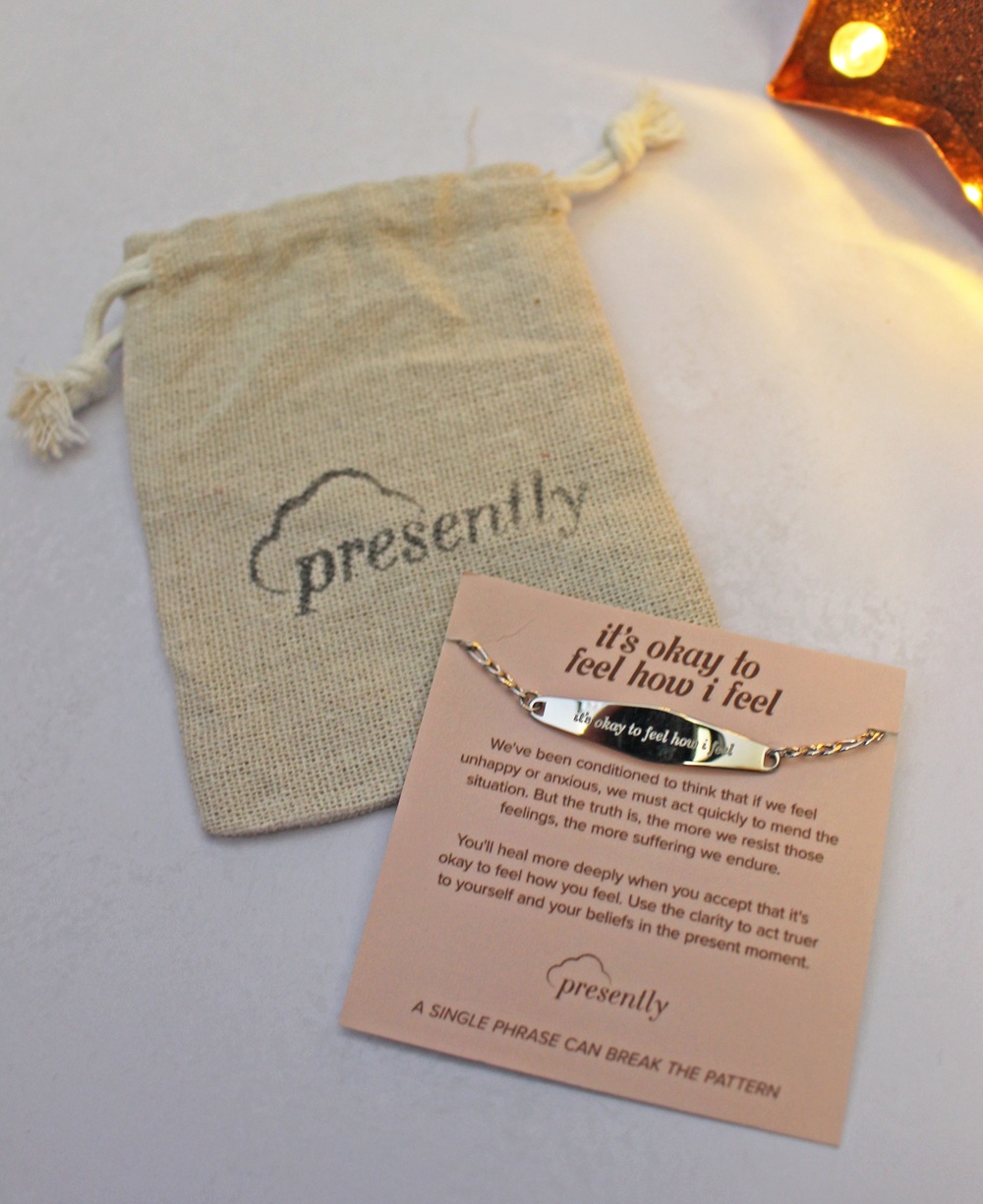 A delicate silver chain bracelet with a bar inscribed it's okay to feel how I feel, displayed on an informational card next to a small cloth drawstring bag with the Presently logo.
