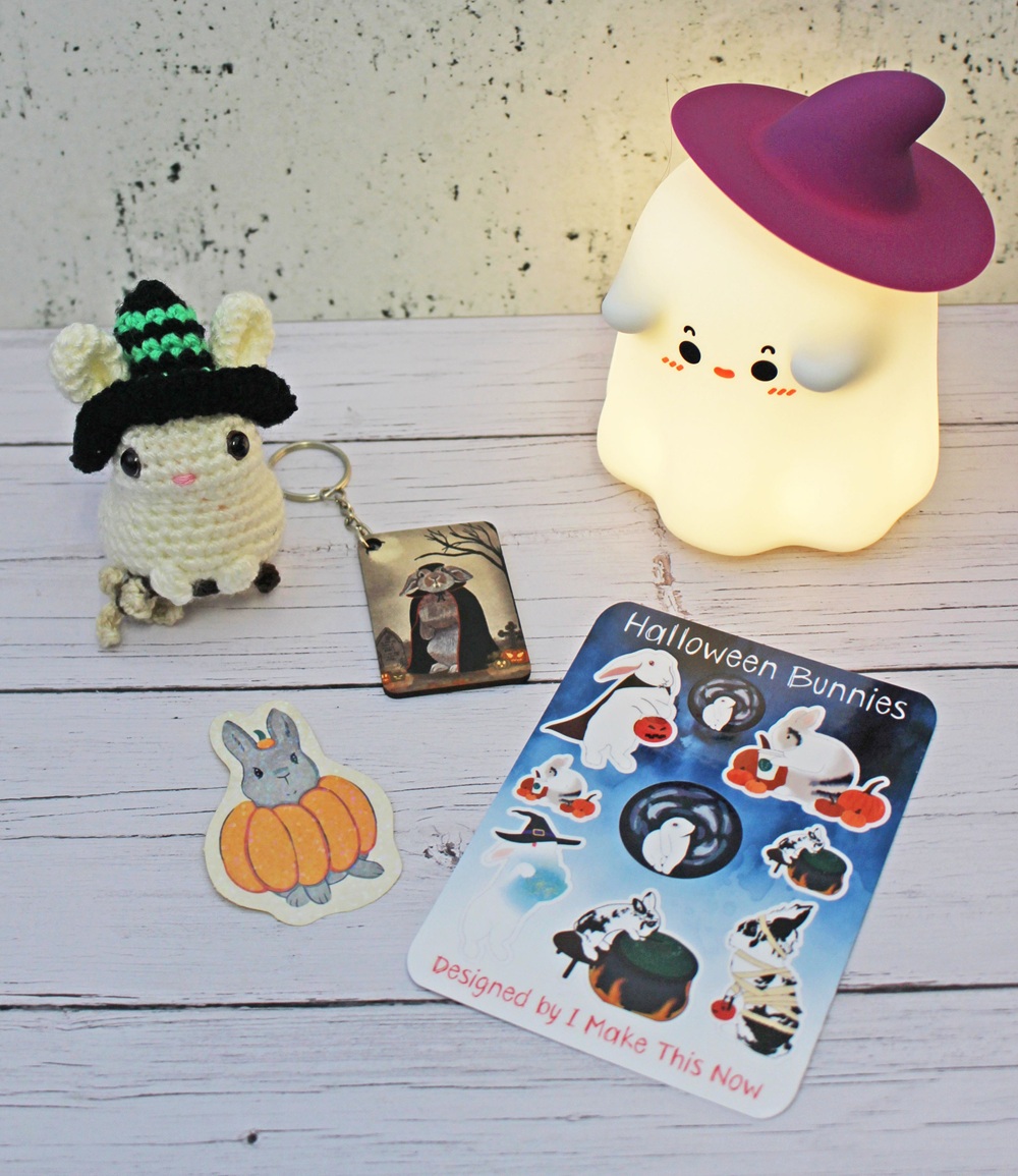 Crochet bunny in a witch hat, a Hallowe’en keyring, a bunny-in-pumpkin sticker, a sticker sheet labelled Hallowe’en Bunnies, and a glowing ghost lamp with a purple hat on a white wood surface.