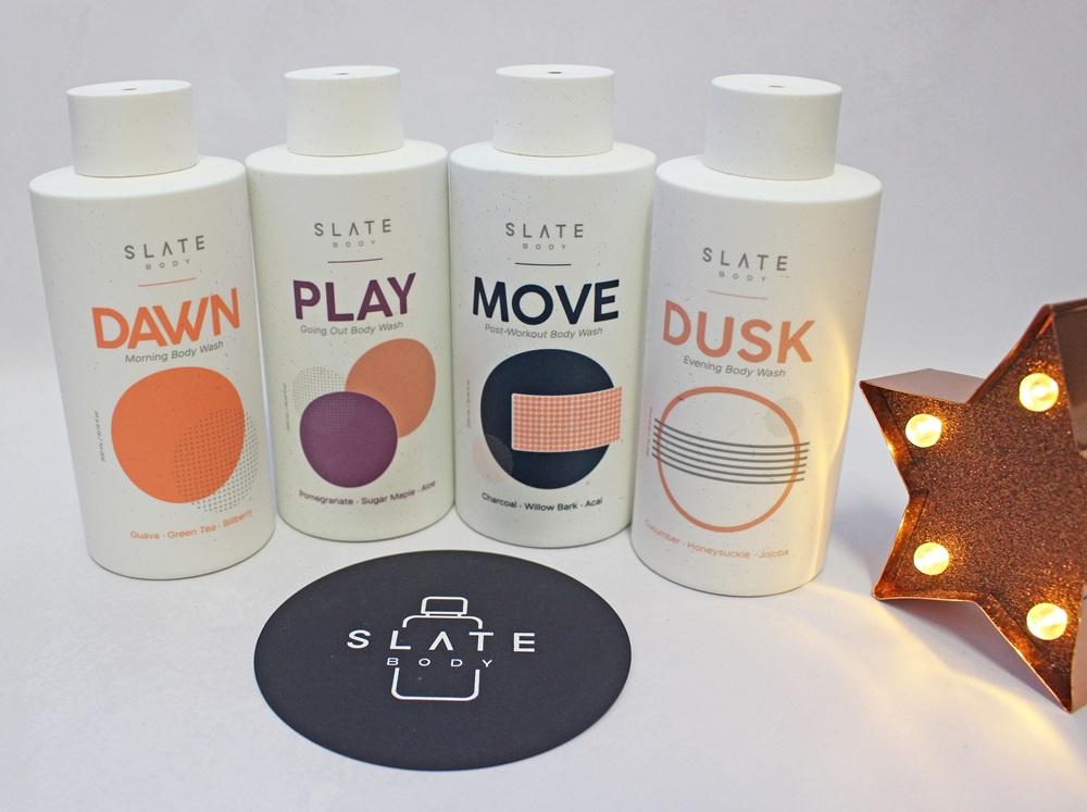 Four bottles of Slate Body wash labelled Dawn, Play, Move, and Dusk, arranged next to a Slate Body logo coaster and a lighted star decoration.