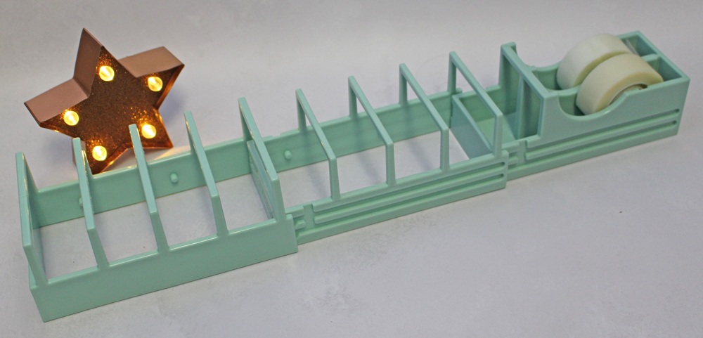 A light green plastic desk organiser with multiple vertical slots and a built-in tape dispenser holding two rolls of tape, next to a small light-up star.