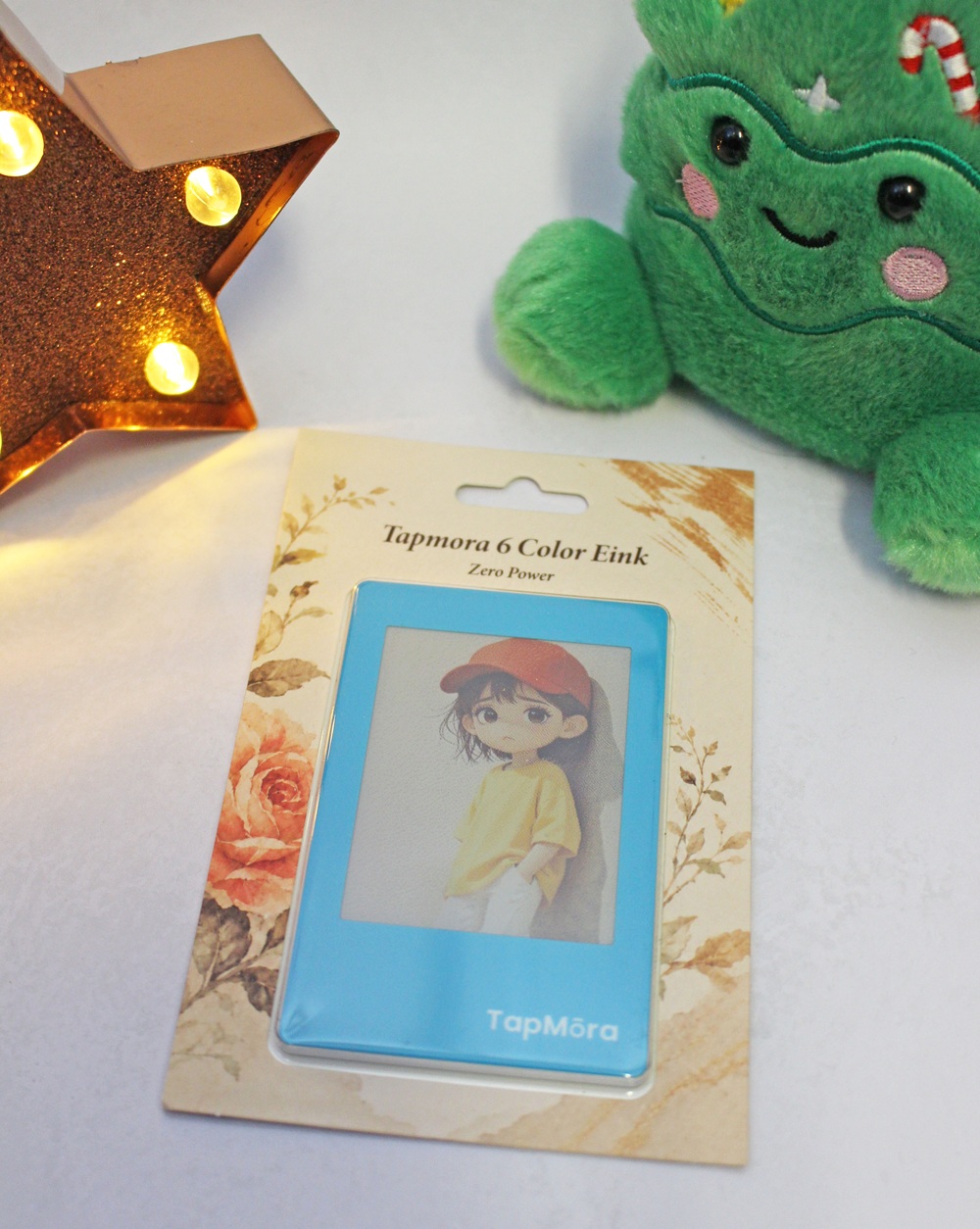 A Tapmora 6 Colour Eink digital display in its packaging, featuring an illustration of a girl in a red cap and yellow shirt, sitting next to a light-up star ornament and a plush green Christmas tree.