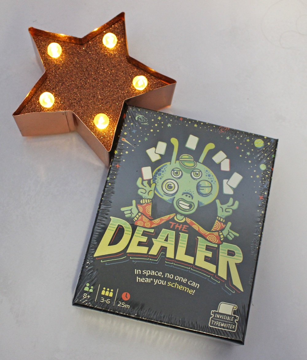 The Dealer card game box featuring a green three-eyed alien illustration, next to a star-shaped marquee light.