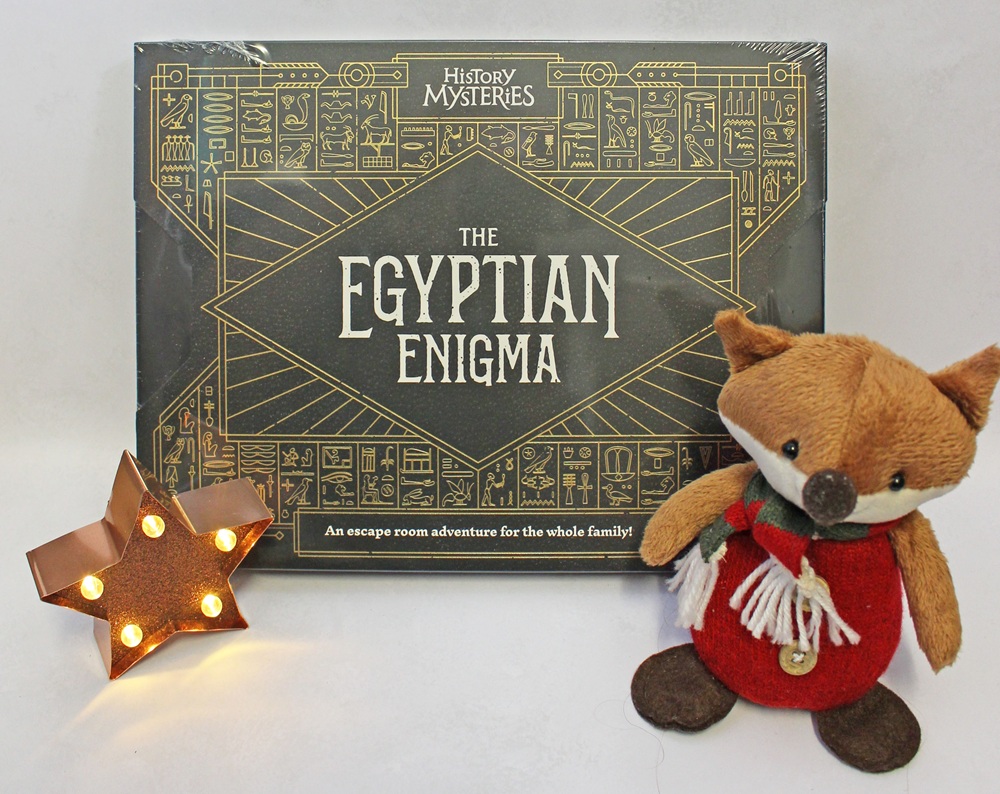 The Egyptian Enigma escape room board game box next to a small plush fox and a lit metal star.