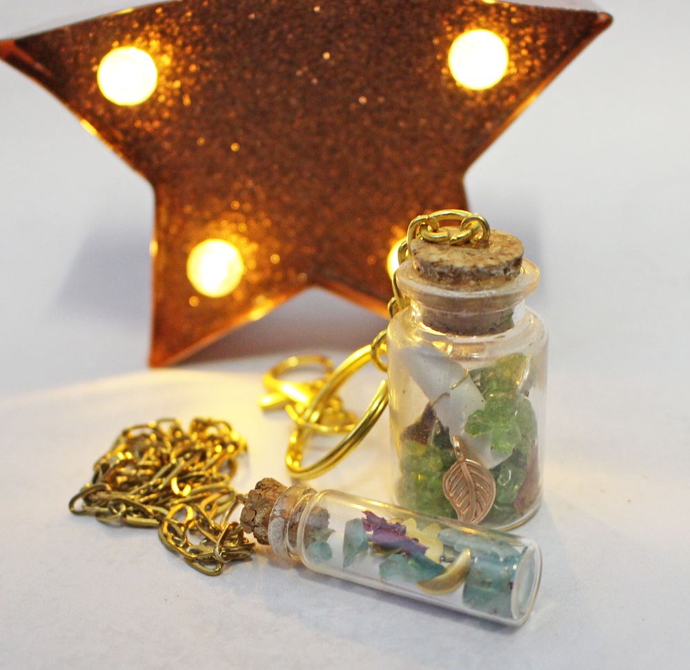 Two small glass bottles with cork stoppers containing crystals and charms, one on a gold keyring and the other on a gold chain, set in front of a lit, star-shaped marquee light.