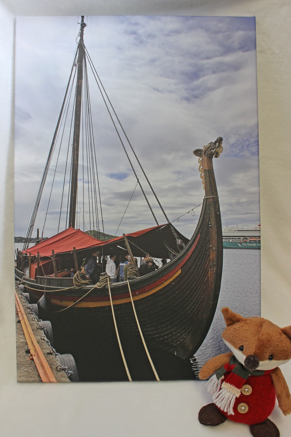 A canvas print of a wooden Viking ship docked at a pier, with a small stuffed fox wearing a scarf and jumper in the foreground.