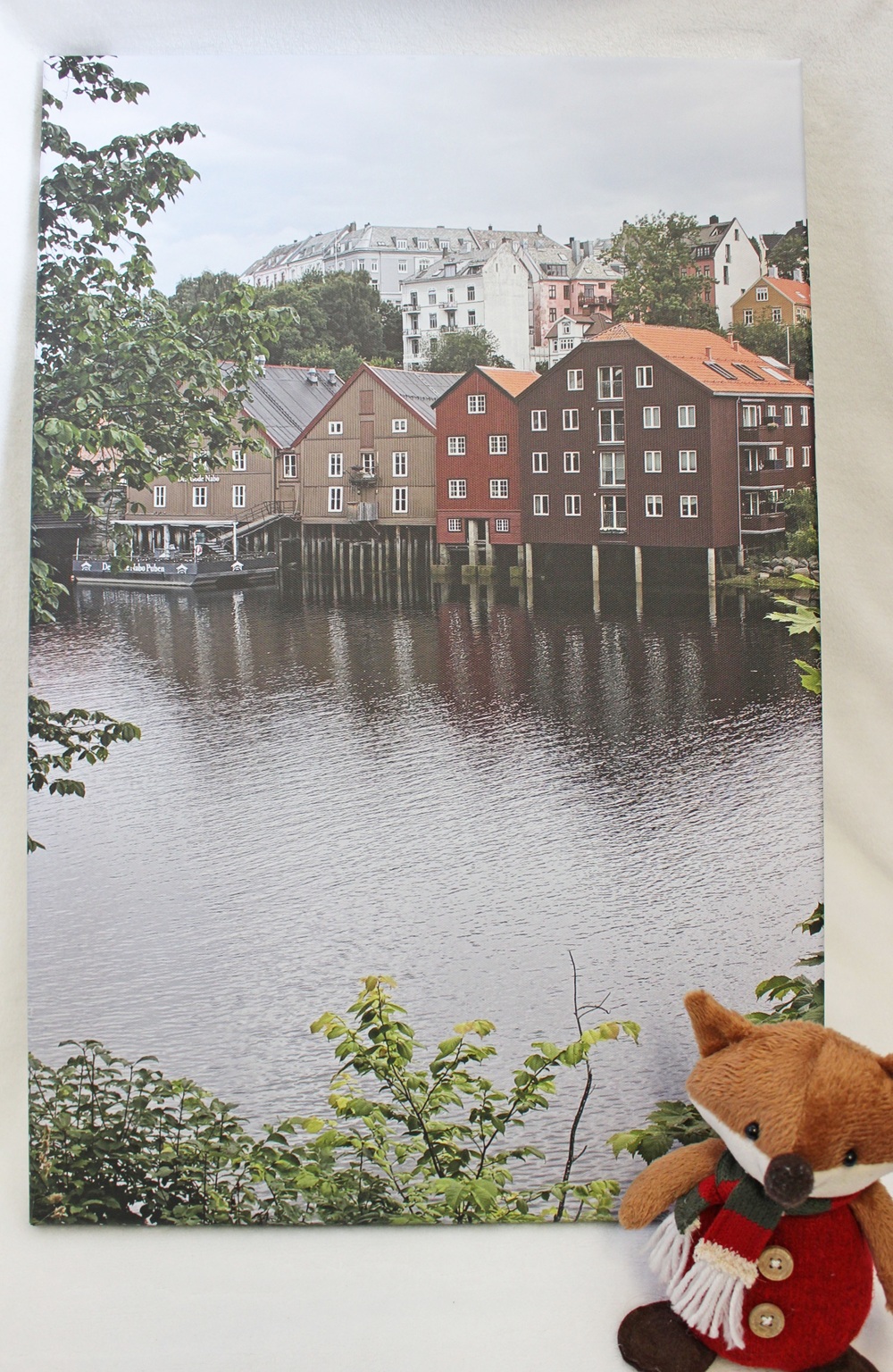 A small plush fox wearing a red coat and scarf sits in the corner of a framed photo showing colourful buildings on stilts over a river in a city.