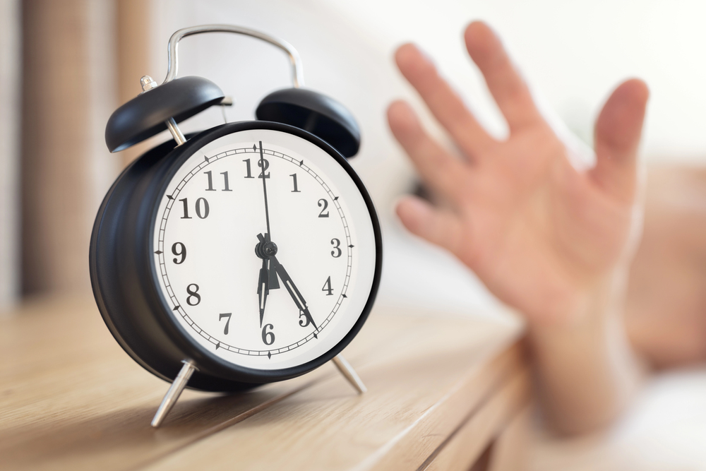 A person reaching out to hit a black analogue alarm clock sitting on a bedside table.