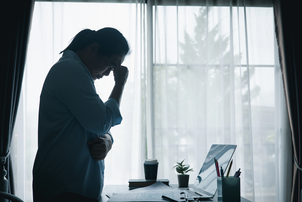 A silhouette of a person standing at a desk in front of a window, holding their head in frustration.