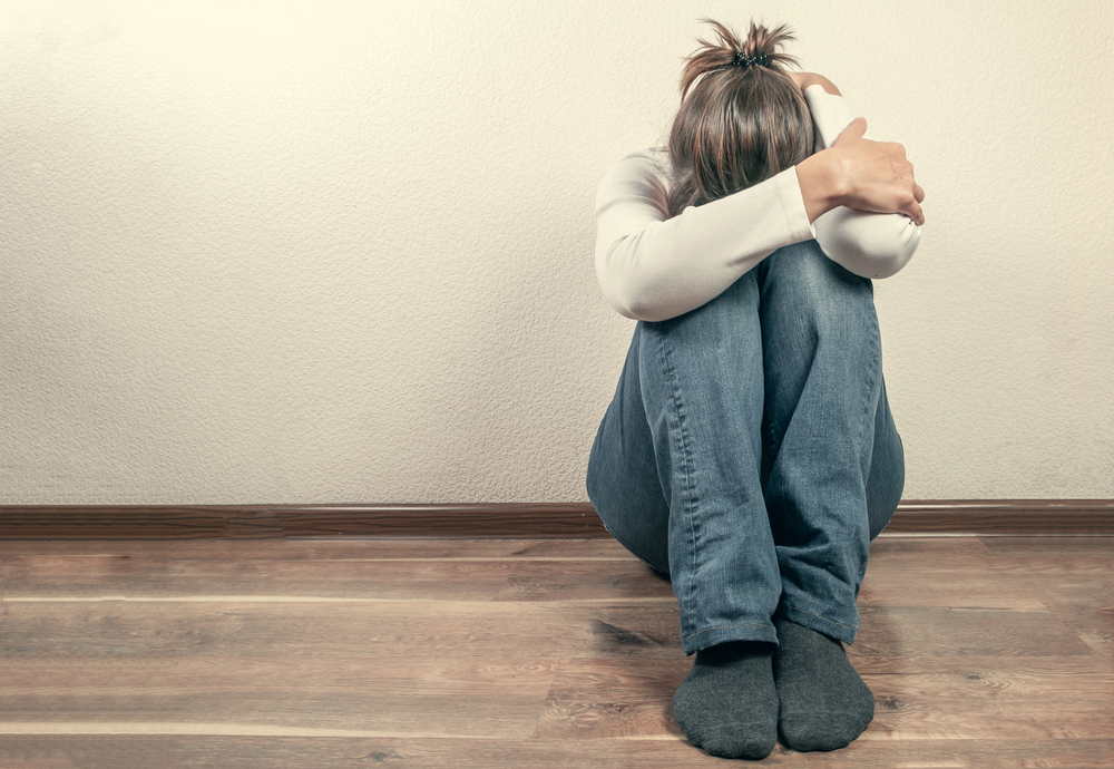 A person sitting on a wooden floor with their head tucked into their arms, knees drawn to their chest.