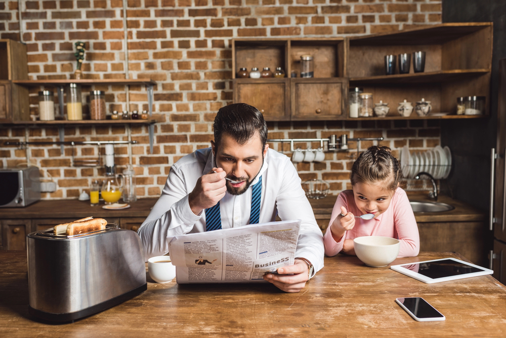A father in a white shirt and tie reads a newspaper whilst eating breakfast next to his young daughter, who is eating cereal at a kitchen table.