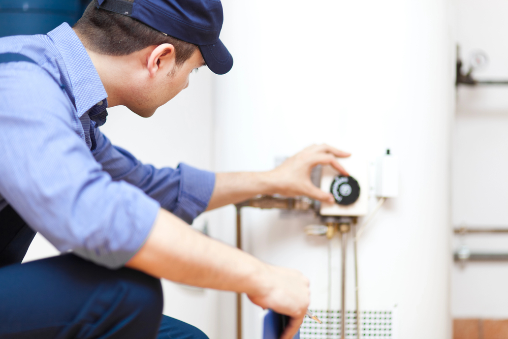 A technician wearing a blue uniform cap adjusting the dial on a water heater.