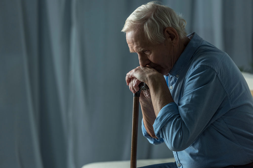 An elderly man with white hair sitting and leaning his chin on his hands, which are resting on the handle of a wooden walking stick.