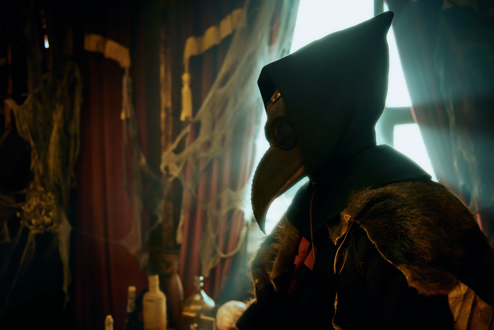 A person wearing a plague doctor mask and hooded cloak stands in a dimly lit, draped room.