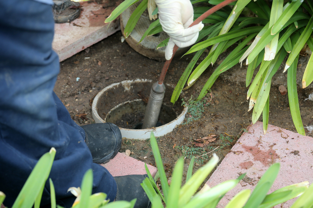 A person wearing a glove uses a plumber's rod to clear an outdoor drainpipe.
